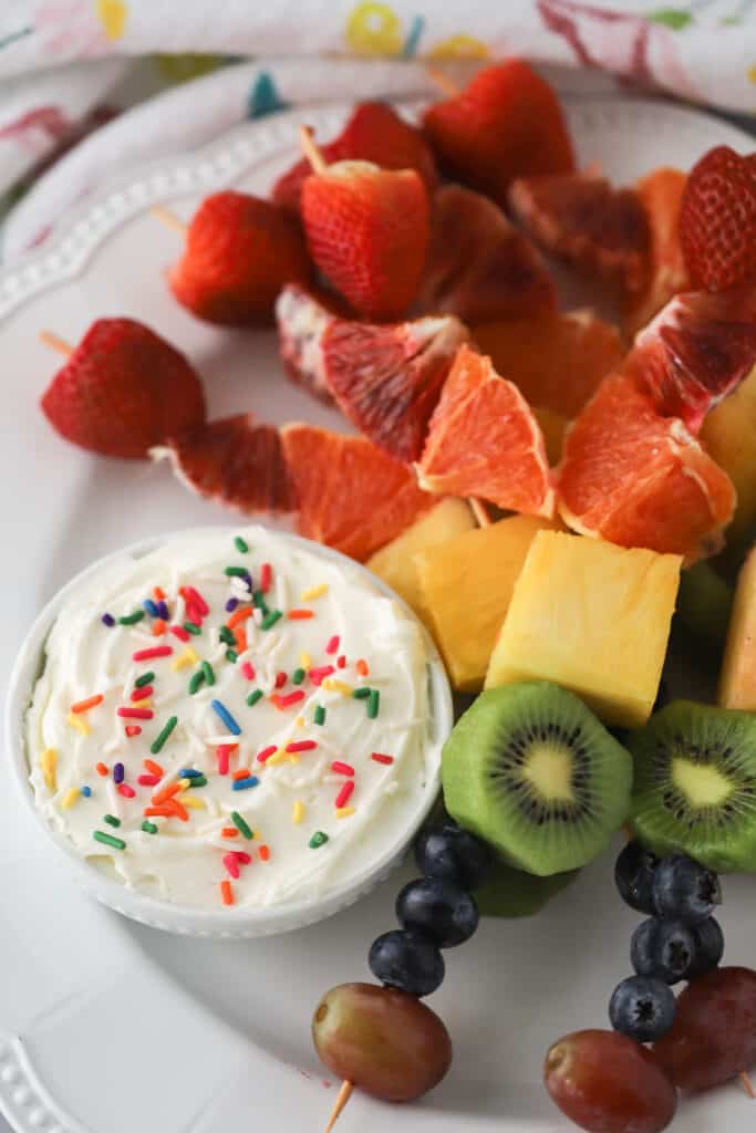 rainbow fruit kebabs on a serving plate with a side of cream cheese fruit dip.