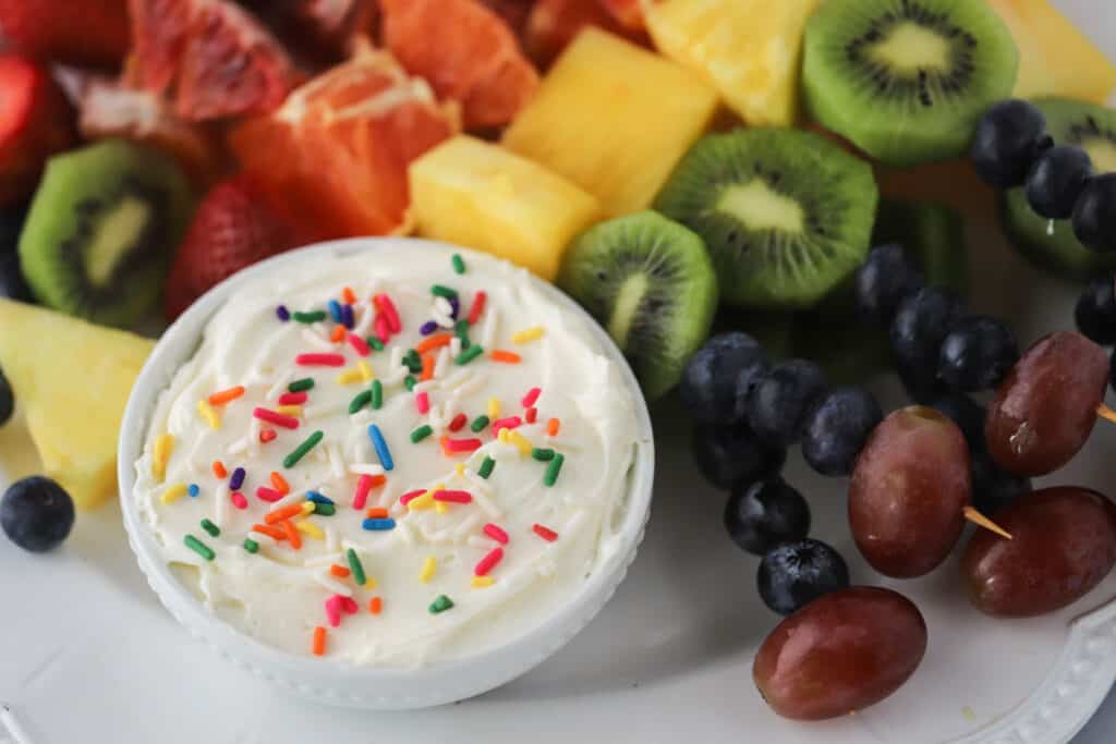 A bowl with cheesecake fruit dip with skewers of fresh fruit.