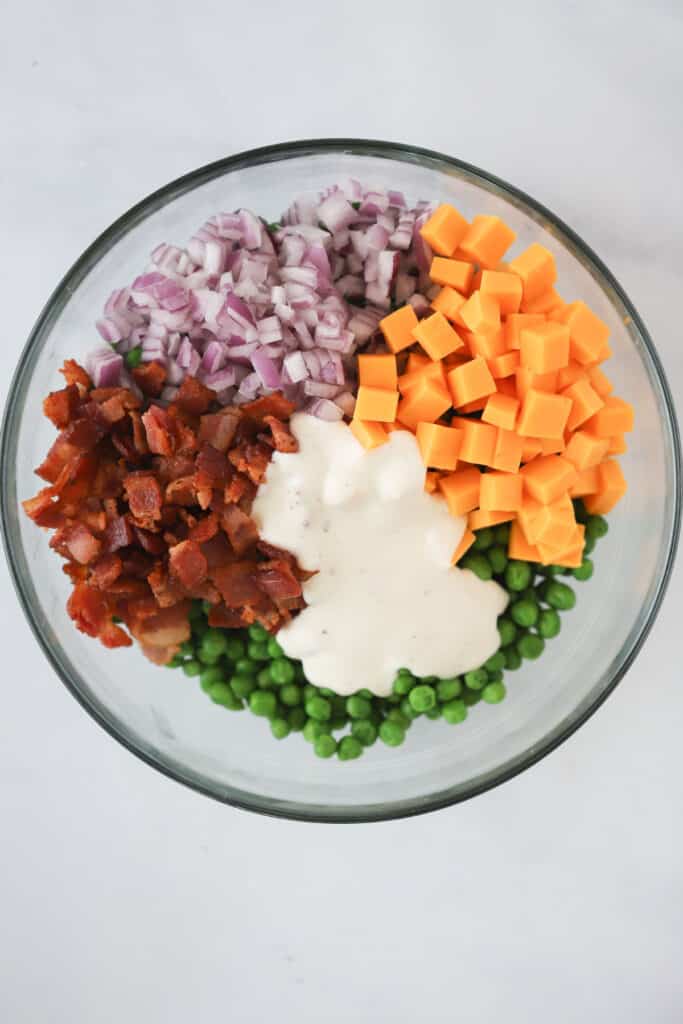 A large glass mixing bowl with ingredients for making this sweet pea recipe including bacon, peas, red onions, ranch dressing, and cheddar cheese.