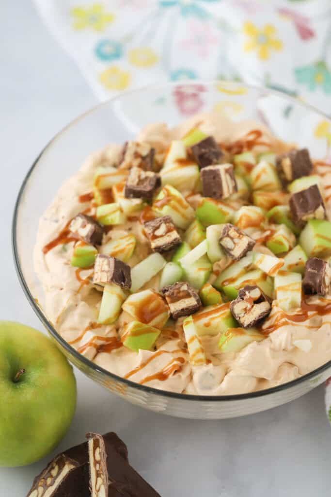 A large bowl full of Snickers Apple Salad topped with caramel sauce, Granny smith apples, and bite sized Snickers candy bars.