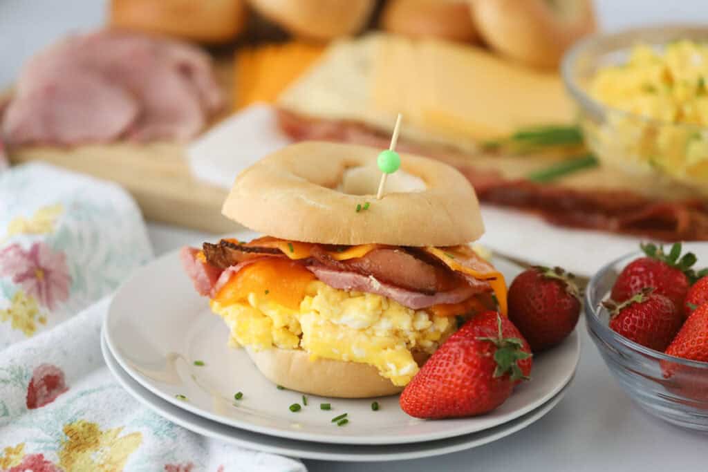 A plate with a loaded up Breakfast bagel sandwich full of scrambled eggs, ham, and bacon.