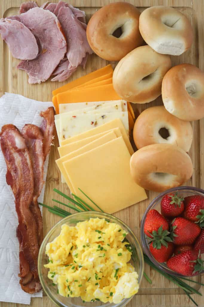 A cutting board full of bagels, ham, bacon, sliced cheese, scrambled eggs, and fresh strawberries for making bagel breakfast sandwiches.