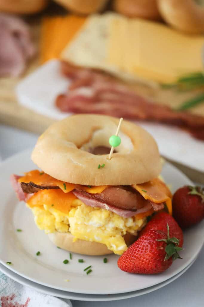 A Bagel Breakfast Sandwich filled with scrambled eggs, bacon, and ham on a plate with fresh strawberries.