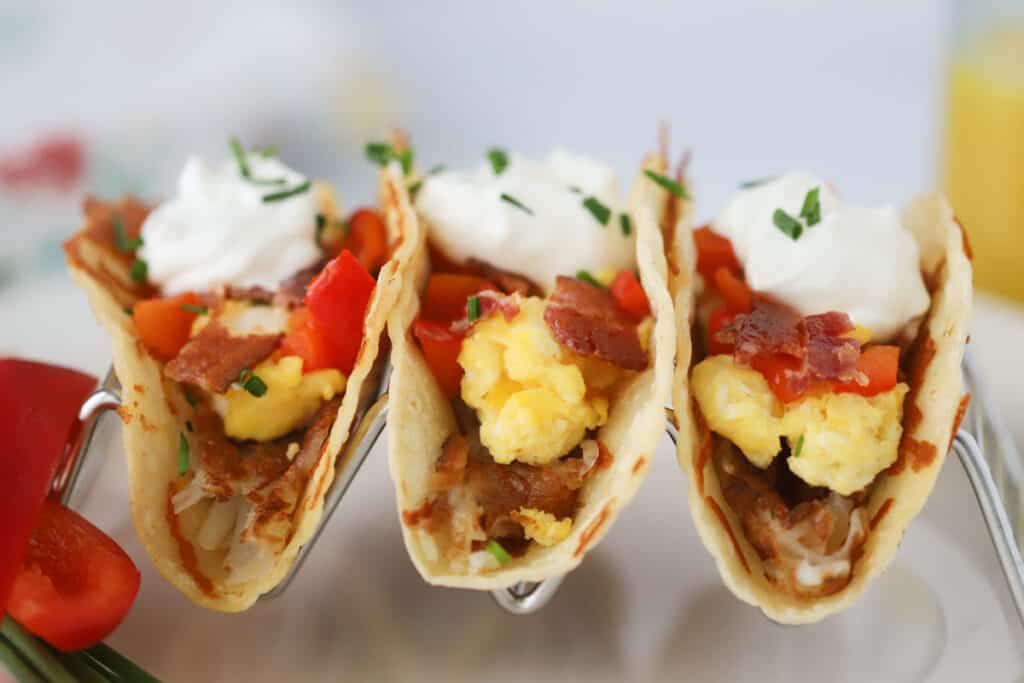 Easy breakfast tacos in a taco holder filled with hash browned potatoes, scrambled eggs, bacon bits, veggies, and topped with sour cream.