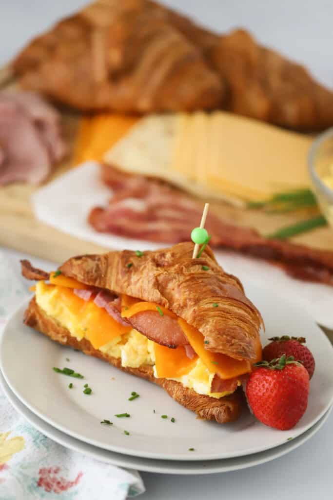 A serving plate with a croissant breakfast sandwich filled with scrambled eggs, melted cheese, and ham.