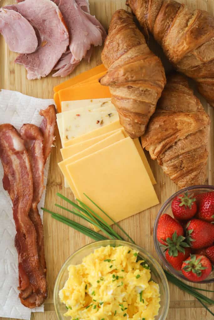 A cutting board with croissants and fillings to make breakfast sandwiches.