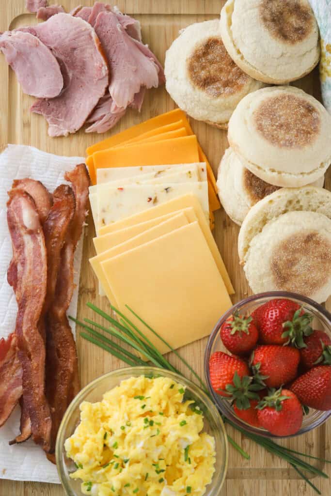 Egg McMuffin ingredients laid out on a cutting board ready to assemble, including sliced cheese, bacon, ham, scrambled eggs, and sliced English muffins.