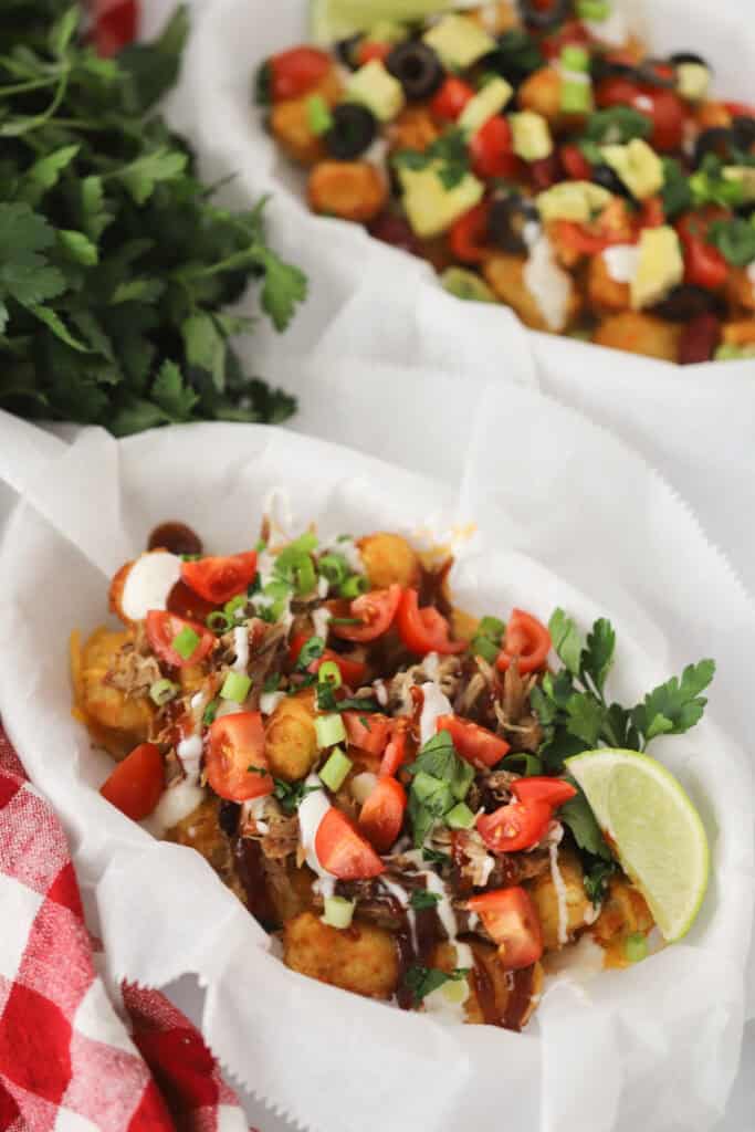A parchment paper lined basket full of tochos made from baking tater tots with pork, cheese, and sauces and then topping with tomatoes, green onions, and cilantro.