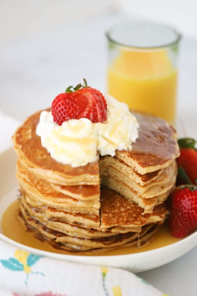 A stack of whole wheat pancakes on a plate with syrup poured over and topped with whipped cream and berries.