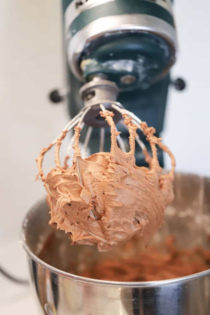 A stand mixer with a whisk attachment and a bowl of creamy chocolate frosting.