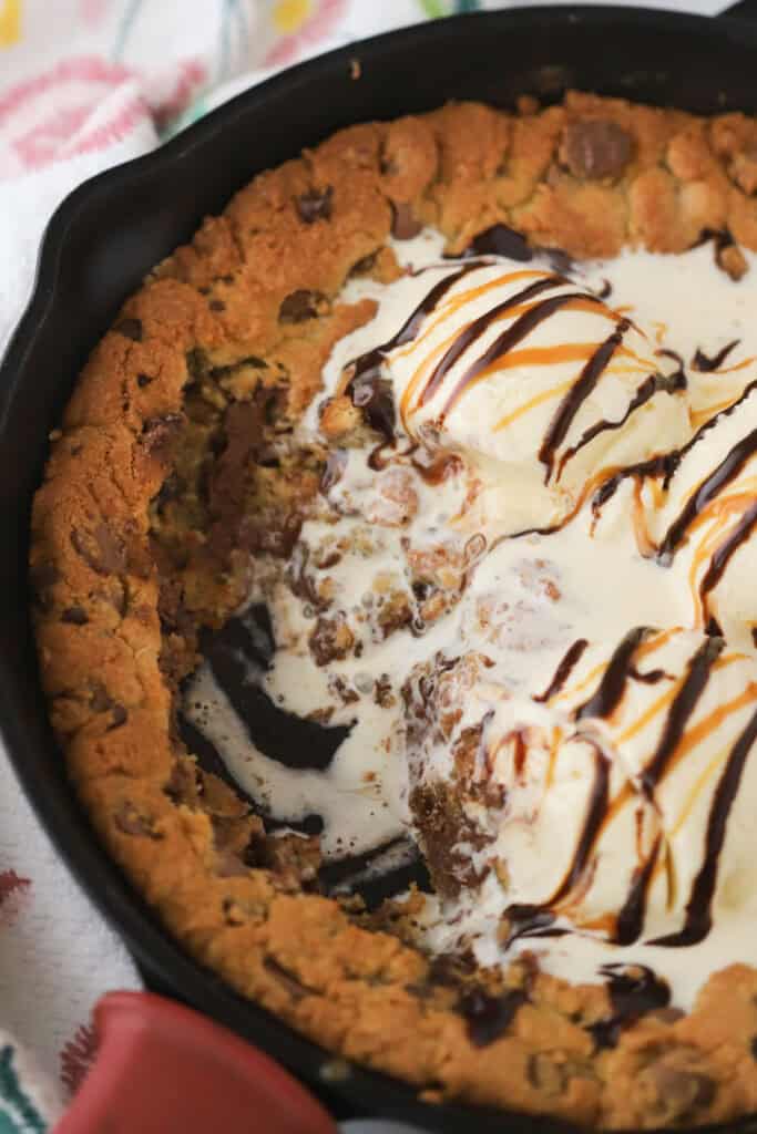 A cast iron skillet with a chocolate chip skillet cookie baked inside and topped with vanilla ice cream, chocolate sauce, and caramel sauce.