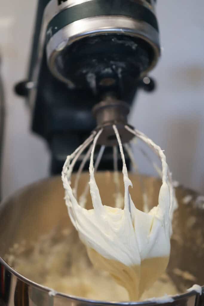 A stand mixer with the whisk attachment and a bowl full of white frosting.