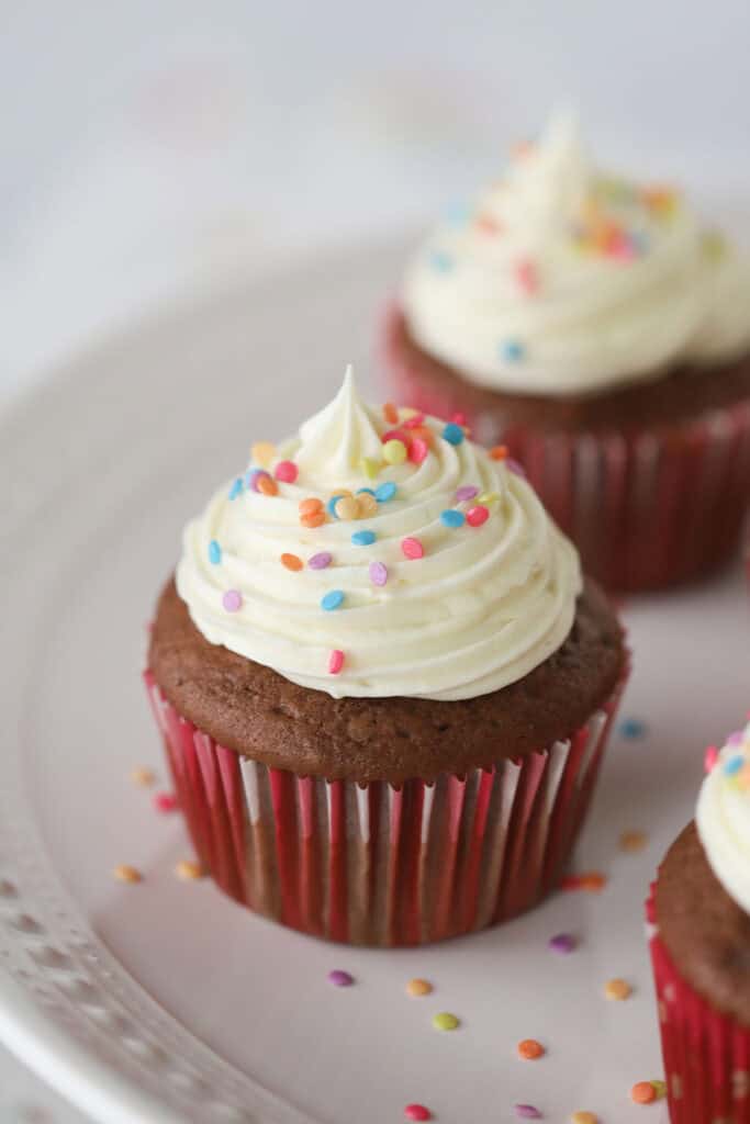 A plate with chocolate cupcakes piped with white chocolate cream cheese frosting, and topped with colored sprinkles.