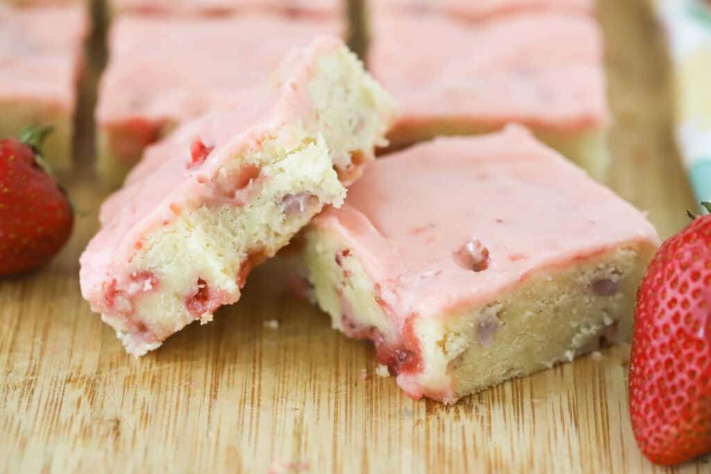 Squares of strawberry lemon blondies topped with pink strawberry icing on a countertop.