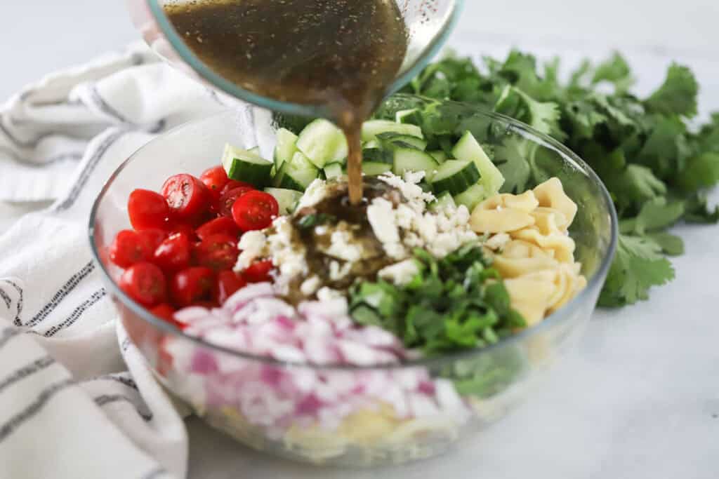 A large bowl with greek vinaigrette being poured over tortellini pasta salad ingredients.