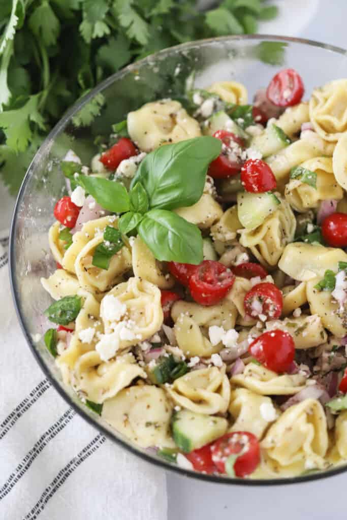 A large glass serving bowl full of greek tortellini salad topped with fresh basil.