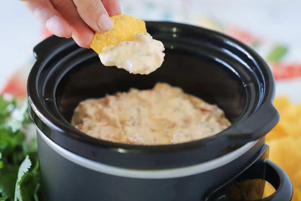 A hand dipping a chip into dip with sausage and cheese that is in a crockpot.