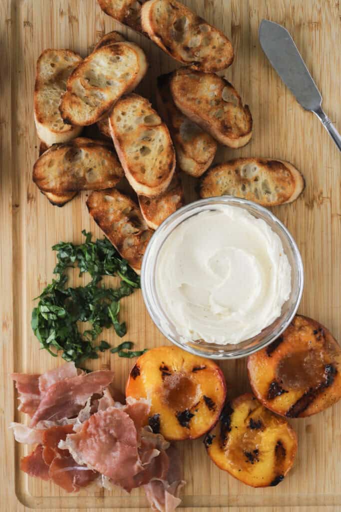 A cutting board with grilled baguettes, whipped honey cream cheese, grilled peach slices, pancetta, and sliced basil for making peach bruschetta.