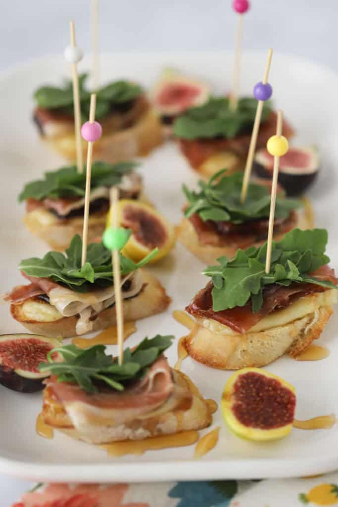 A white serving plate with fig and brie crostini stuck with decorative toothpicks.