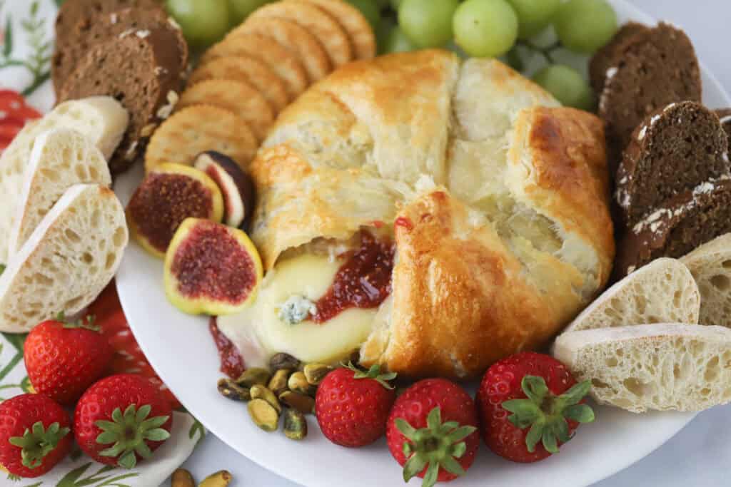 a serving plate with baked brie with fig jam wrapped in puff pastry, surrounded by fresh fruit, sliced bread, and crackers.