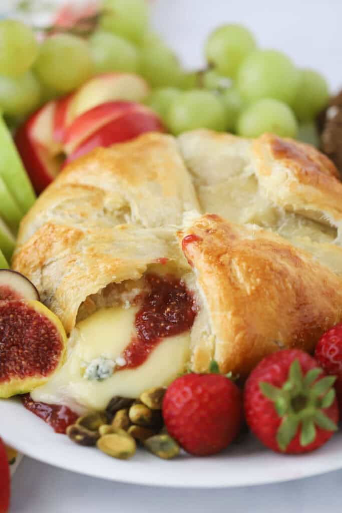 Baked brie with fig jam, gorgonzola cheese, honey, and rosemary all surrounded by fresh fruit on a serving plate.