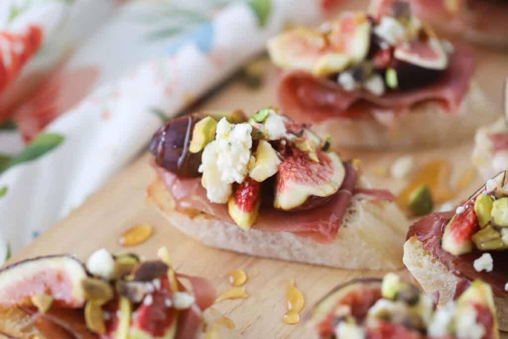 A serving platter with prosciutto and fig crostini topped with honey, cheese, and pistachios.