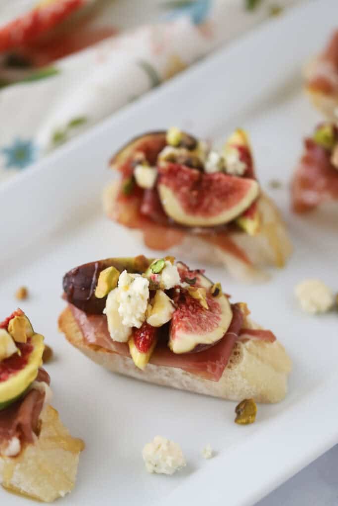 A white serving plate with fig and prosciutto crostini topped with gorgonzola and pistachios.