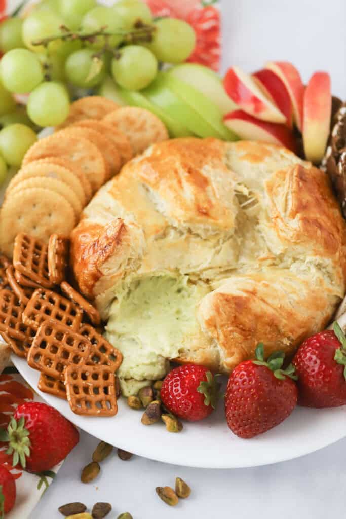 A round of stuffed Parmesan and Pesto Puff Pastry, surrounded by crackers, pretzels, and fresh fruit.