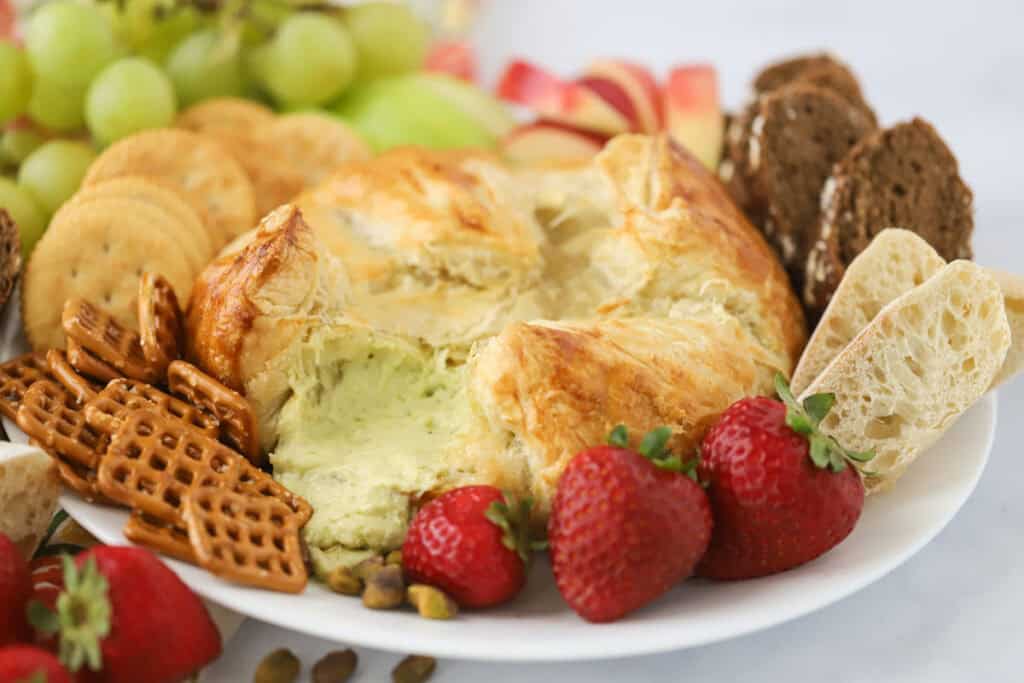 A baked puff pastry round filled with cheese and pesto on a plate with crackers, fruit, sliced, bread, and pretzels.