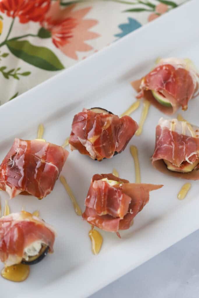 A white serving plate with rows of prosciutto wrapped figs topped with honey.