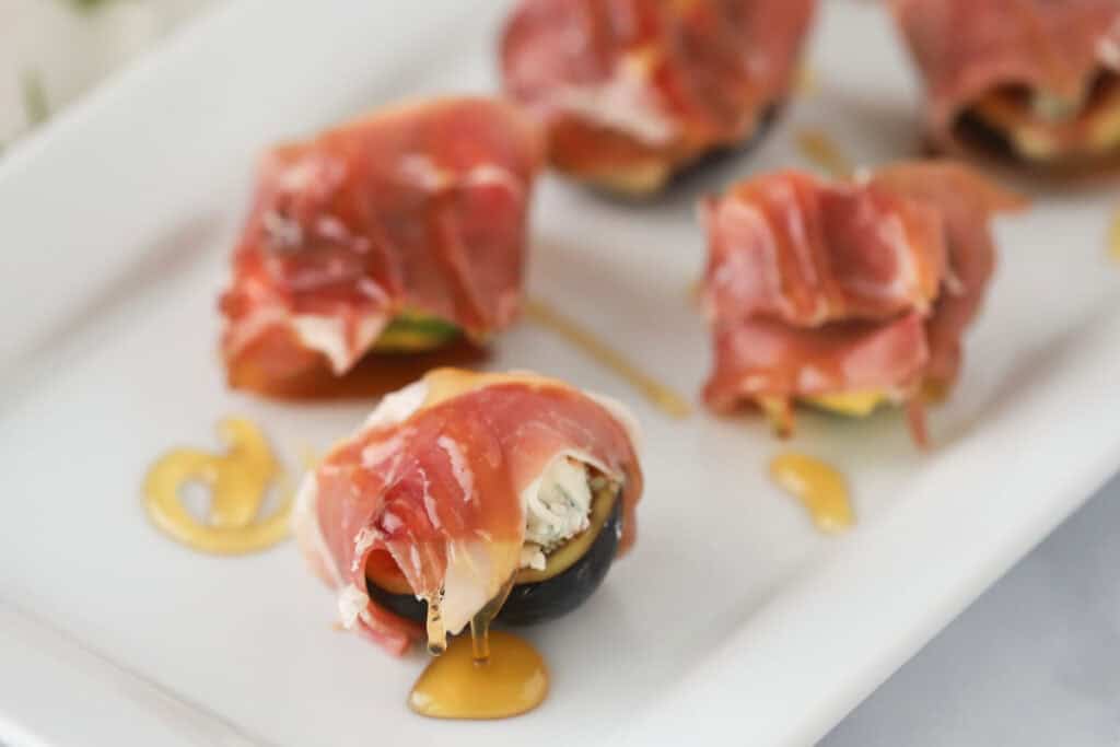 A serving plate full of figs wrapped in prosciutto and topped with honey.