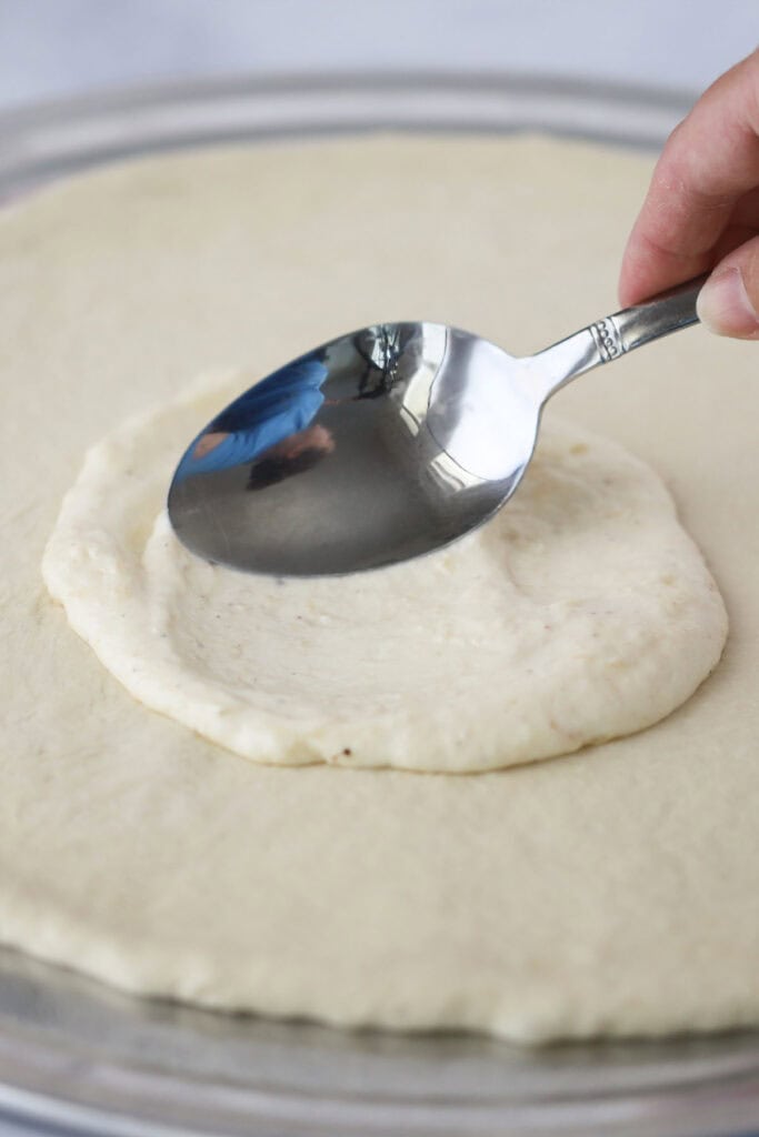 A spoon spreading this white sauce recipe for pizza over pizza dough.