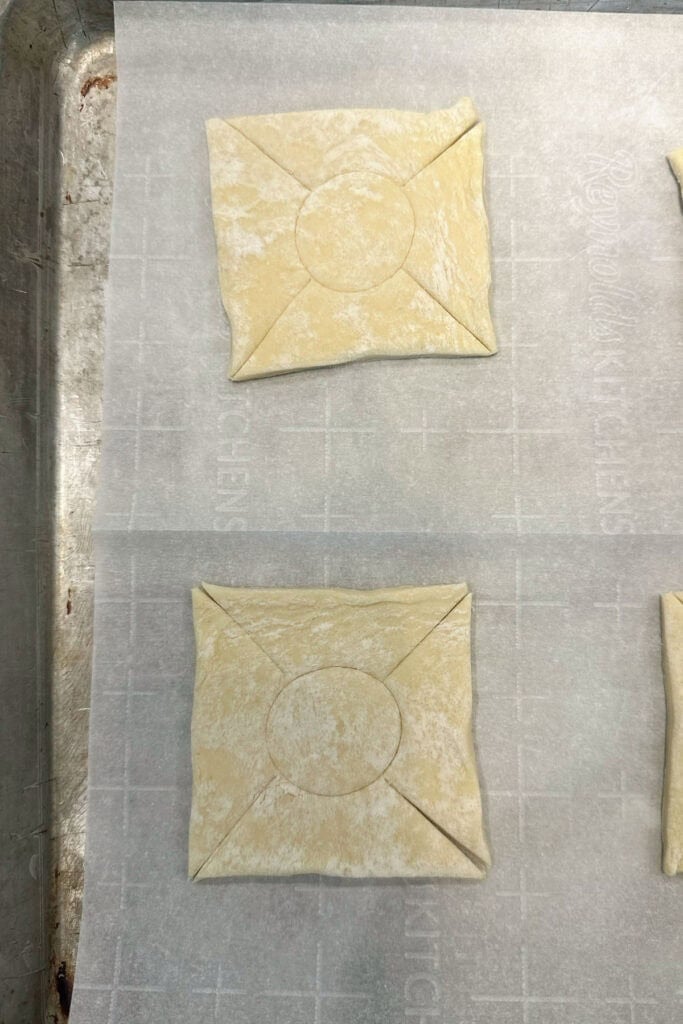 Puff pastry dough on a baking sheet with a circle in the center and cuts on each corner, preparing to fill and bake cheese Danish.