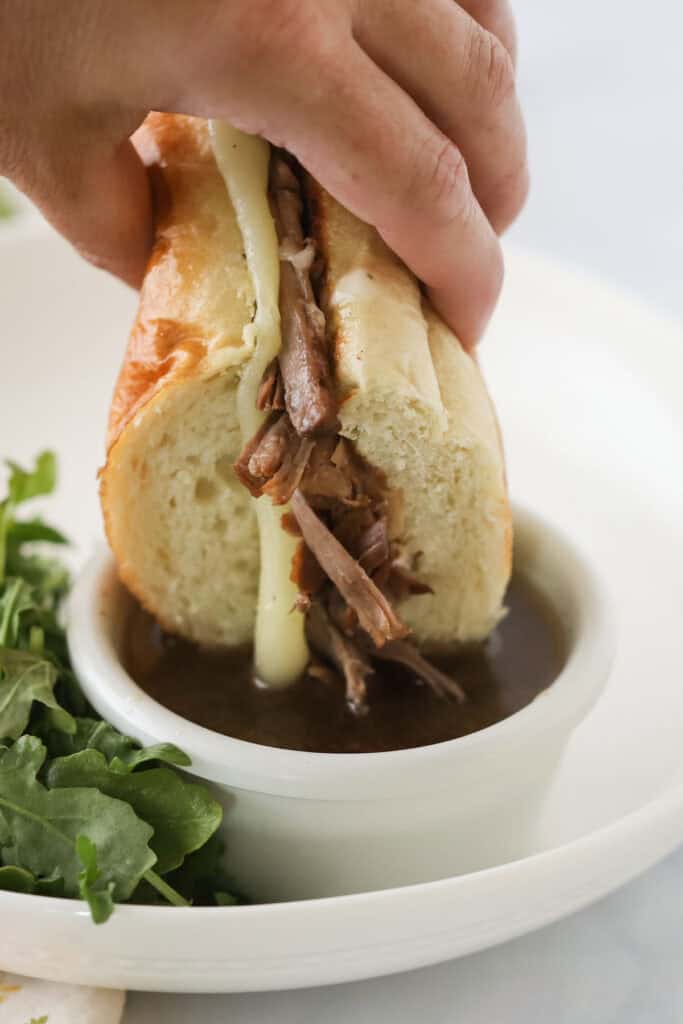 A hand dunking a French Dip sandwich into au jus sauce.