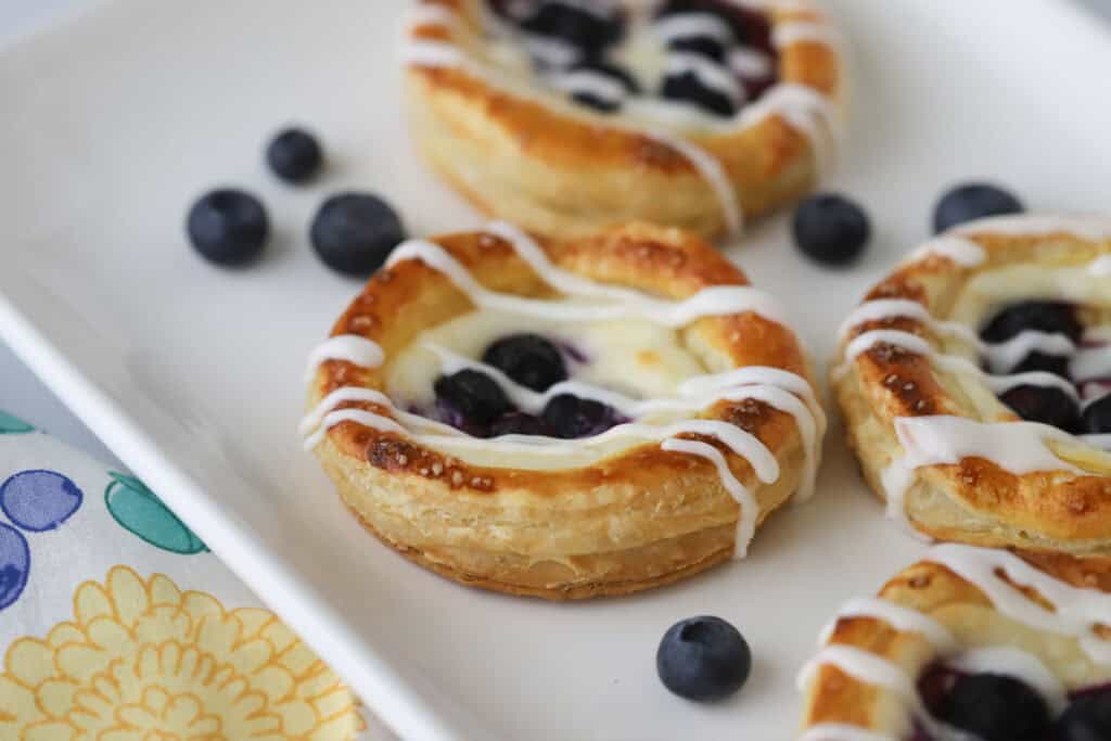 A white plate with freshly baked Blueberry Cheese Danish topped with a drizzle of glaze.