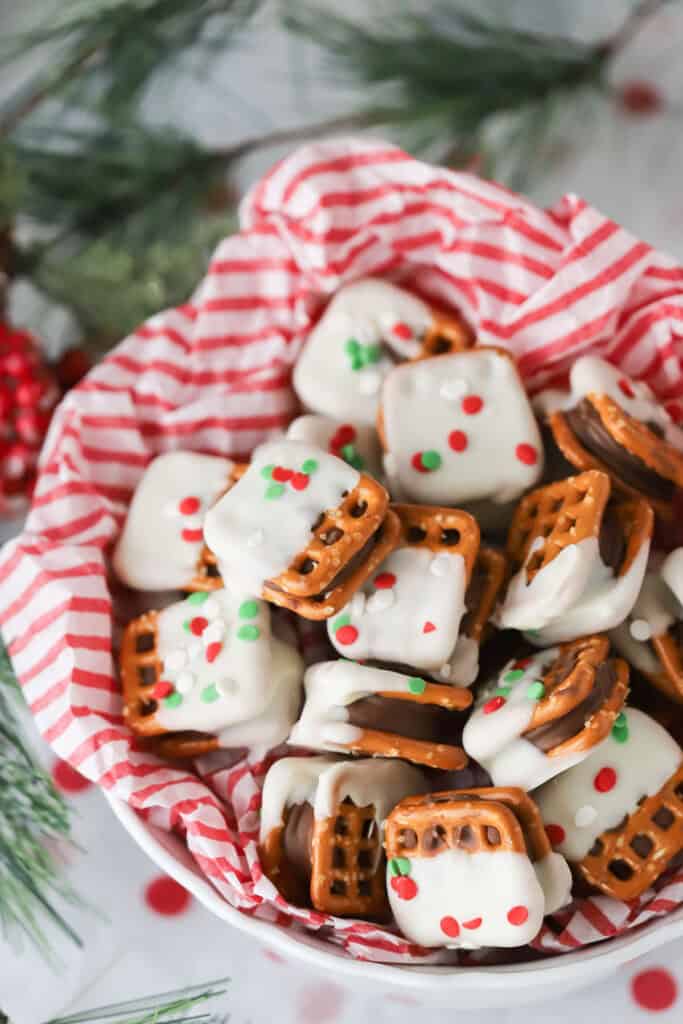 A basket with red and white striped tissue paper, full of pretzel and Rolo treats that have been dipped in white chocolate and covered with red and green sprinkles.