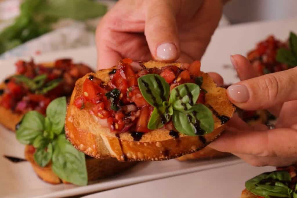 bruschetta appetizer in hands, with a fresh sprig of basil and a drizzle of balsamic glaze. 