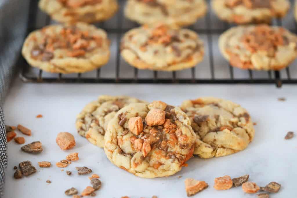A stack of cookies made using this butterfinger cookie recipe, with some cooling on a wire rack behind.