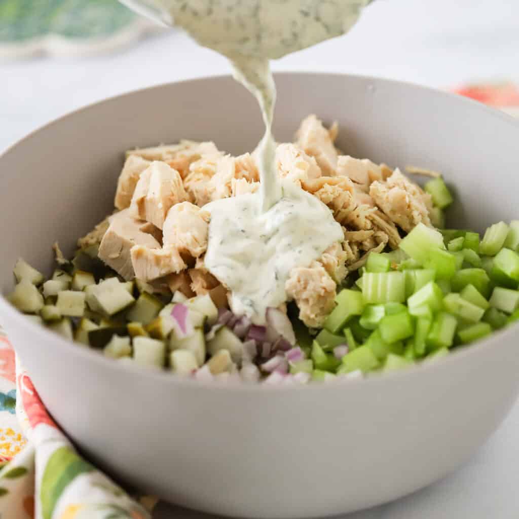 A large white bowl full of chicken salad ingredients with dill pickle dressing being poured over the top.