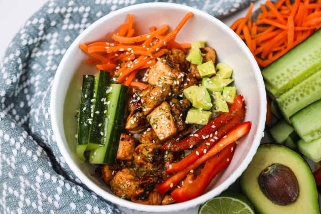 A salmon and rice bowl on a table filled with fresh sliced veggies, avocado, and topped with sesame seeds.