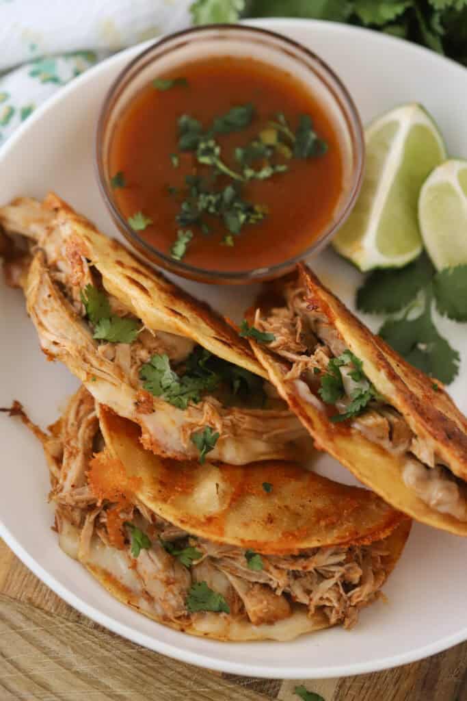 Pork birria tacos on a plate with fresh limes and a side of sauce.