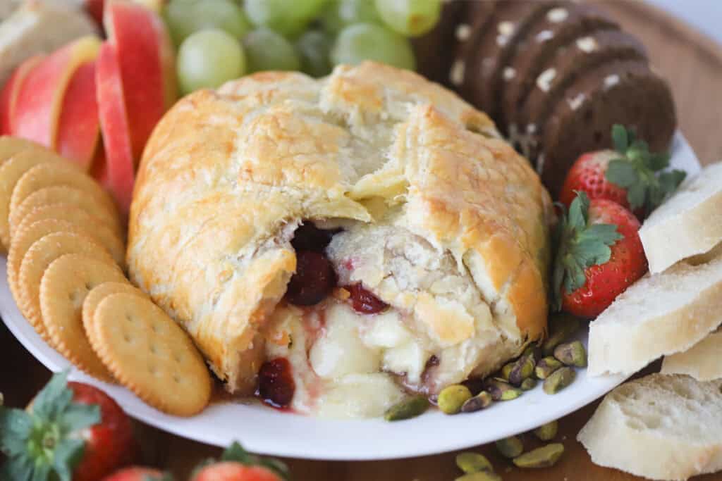 Cranberry baked brie puff pastry on a serving platter surrounded by crackers, bread, and fruit for dipping.