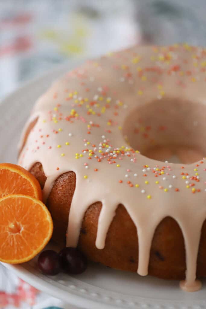 A full cranberry orange bundt cake on a plate, topped with orange frosting and sprinkles.