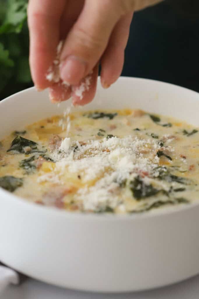 A hand sprinkling Parmesan cheese over the top of Zuppa Toscana in a white bowl.