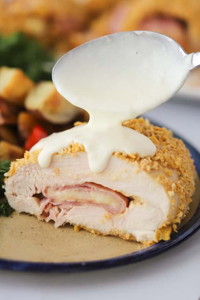 A plate with a slice of chicken cordon bleu with Cornflake breading and a cream sauce being poured over the top.