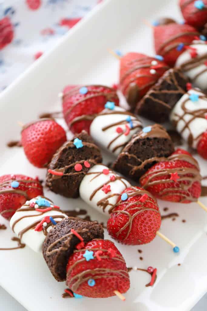 A white serving platter full of strawberry and brownie dessert kabobs, drizzled with chocolate and topped with sprinkles.