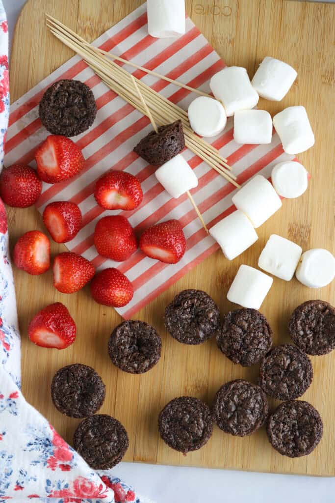 A tabletop with fresh strawberries, brownie bites, large marshmallows, and small skewers.