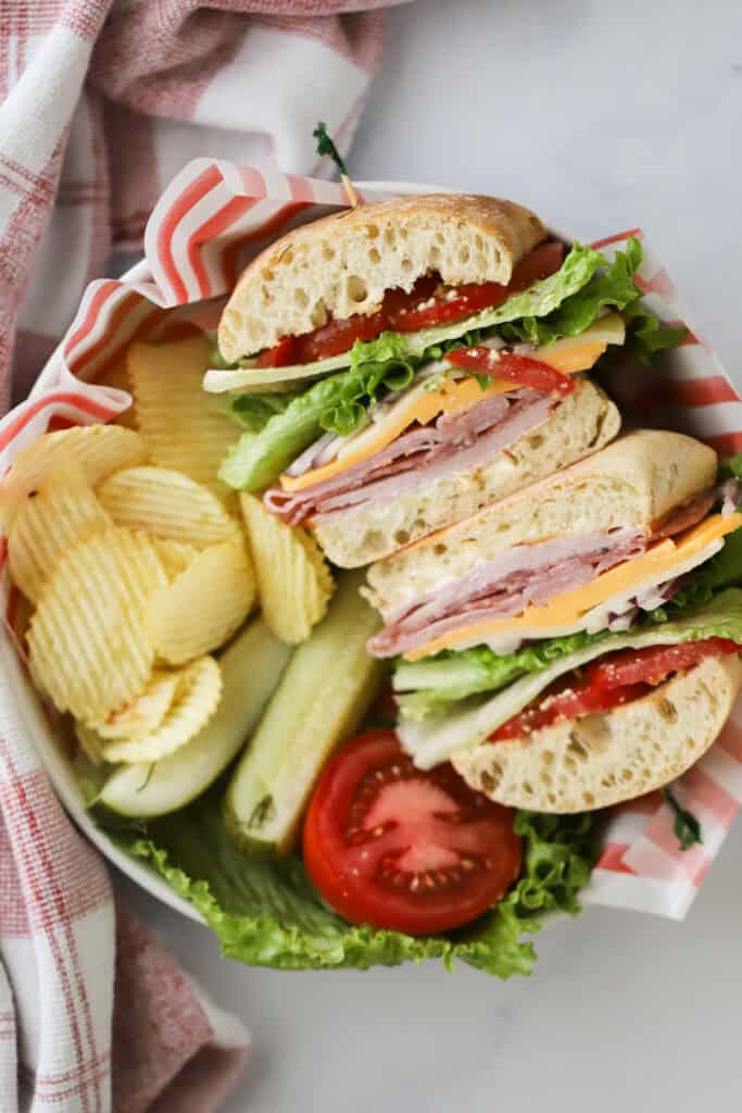 An Italian Club Sandwich in a bowl with chips, pickles, and tomatoes.