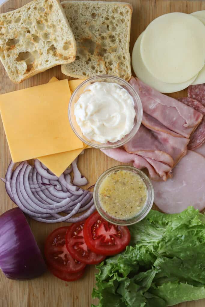 A cutting board full of the ingredients to make an Italian sub, such as Genoa salami, ham, pepperoni, mayo, cheese, onions, tomatoes, lettuce, and toasted bread.