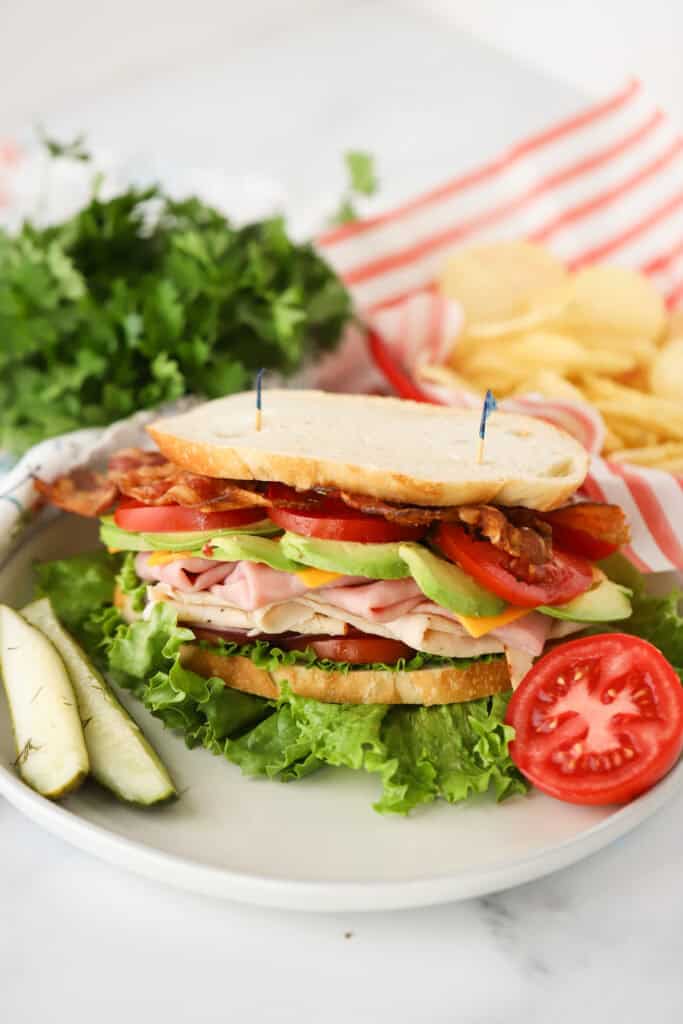 A white dinner plate with a turkey club sandwich and a side of pickles.